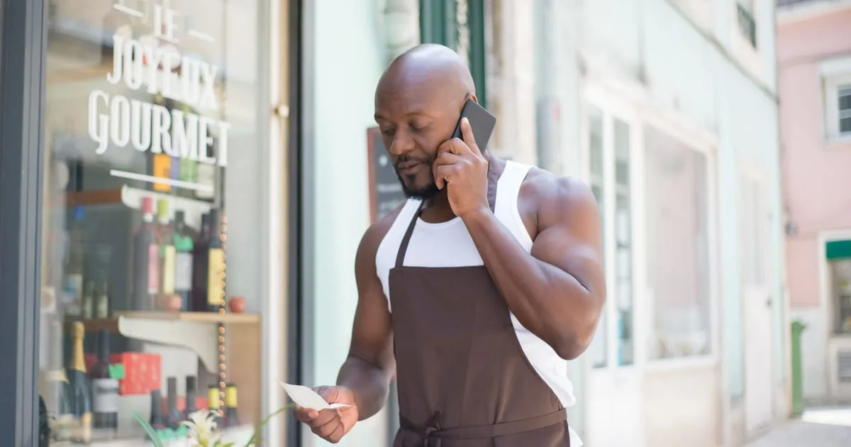 Shop owner standing at the entrance of a small storefront, checking a map listing on a smartphone